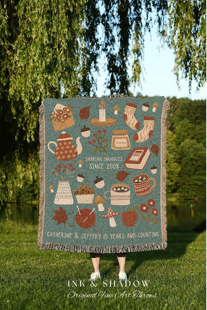 May include: A woven throw blanket with a teal background and a variety of autumn-themed illustrations, including a teapot, a candle, a jar of honey, a book, a cupcake, mushrooms, a mug of hot chocolate, and a ball of yarn. The text on the blanket reads 'Sharing Snuggles Since 2008' and 'Catherine & Jeffery 15 Years and Counting.' The blanket is made by Ink & Shadow Original Fine Art Throws.