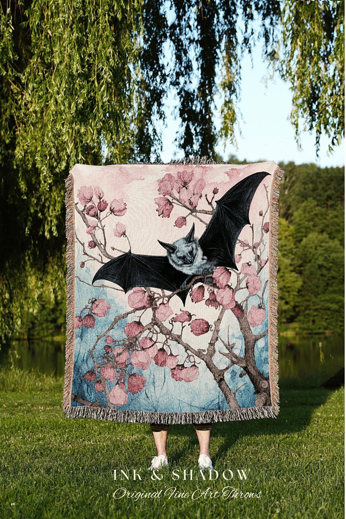 May include: A woven tapestry throw blanket with a pink, blue, and white color scheme. The blanket features a black bat perched on a branch of a flowering tree. The text 'INK & SHADOW Original Fine Art Throws' is printed below the blanket.