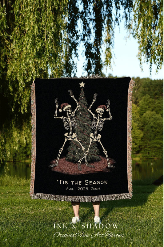 May include: Black woven throw blanket featuring a festive design with two skeleton figures wearing Santa hats, dancing around a Christmas tree. The text "'Tis the Season" is displayed, along with the names Alex and Jamie and the year 2023.