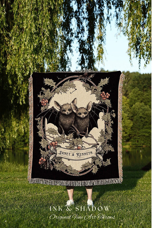 May include: A black woven throw blanket with a cream colored oval frame. The frame contains an illustration of two bats with wings spread. The bats are perched on a branch with holly leaves and berries. A cream colored banner below the bats reads 'Janet & Kenneth'. The blanket has a fringe border.