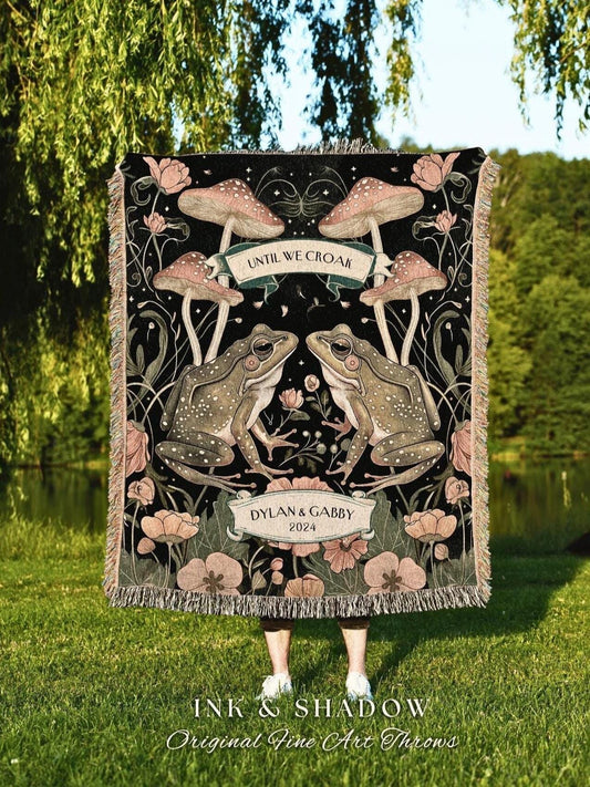 May include: A black throw blanket featuring a whimsical design with two frogs, mushrooms, and floral elements. The blanket has the text "UNTIL WE CROAK" and "DYLAN & GABBY 2024". The blanket is fringed and has the text "INK & SHADOW Original Fine Art Throws".