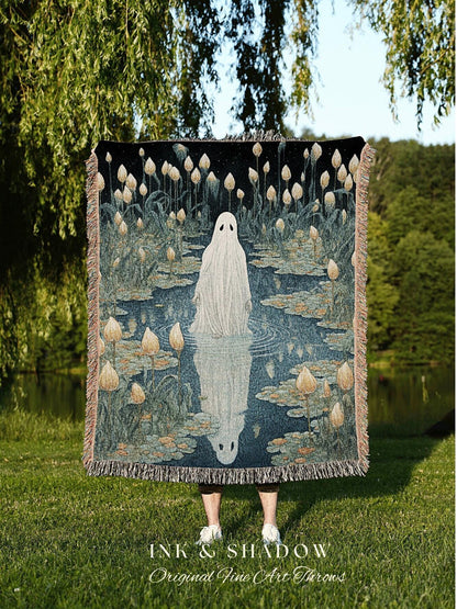 May include: A woven throw blanket featuring a ghostly figure standing in a pond with lily pads and flowers. The artwork is in shades of blue, beige, and black. The blanket has a fringed edge and the text "INK & SHADOW Original Fine Art Throws".