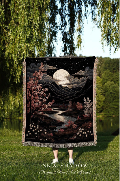 May include: A black throw blanket with a landscape design featuring a full moon, mountains, and a river. The artwork includes pink and white flowering trees. The blanket has a fringed edge and the text "INK & SHADOW Original Fine Art Throws".