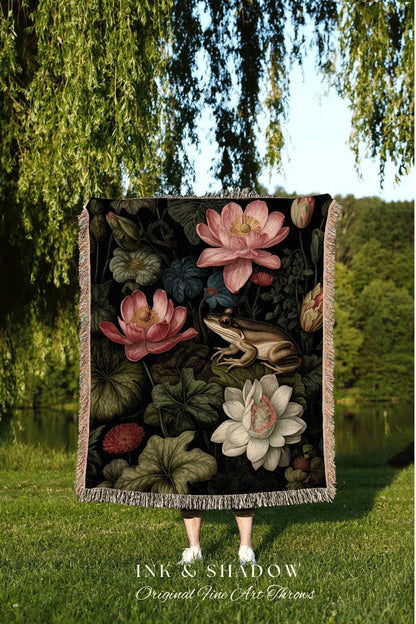 May include: A black throw blanket featuring a botanical design with pink lotus flowers, green lily pads, and a frog. The blanket has a fringed edge and the text "INK & SHADOW Original Fine Art Throws".