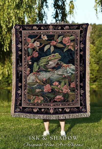 May include: A tapestry throw blanket featuring a detailed illustration of a green frog on a rock surrounded by pink flowers and foliage. The blanket has a dark blue background with a decorative border and fringe. The text "INK & SHADOW Original Fine Art Throws" is at the bottom.