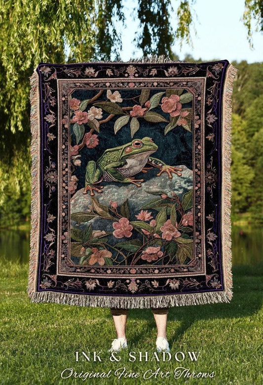 May include: A tapestry throw blanket featuring a detailed illustration of a green frog on a rock surrounded by pink flowers and foliage. The blanket has a dark blue background with a decorative border and fringe. The text "INK & SHADOW Original Fine Art Throws" is at the bottom.