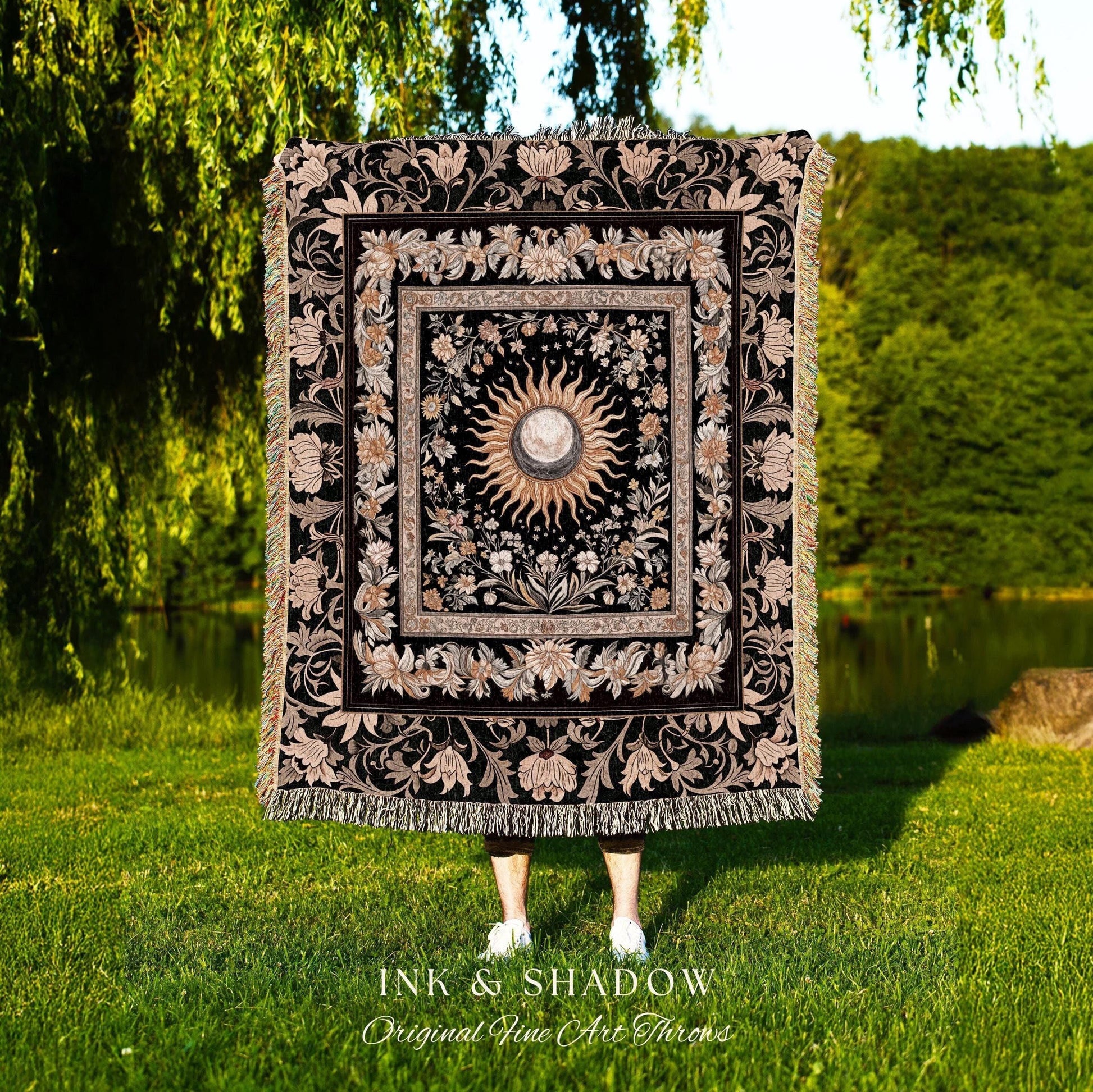 May include: A black woven throw blanket with a gold and white sun and moon design in the center. The design is surrounded by a floral pattern. The blanket has fringe on all sides. The text 'INK & SHADOW Original Fine Art Throws' is at the bottom of the blanket.