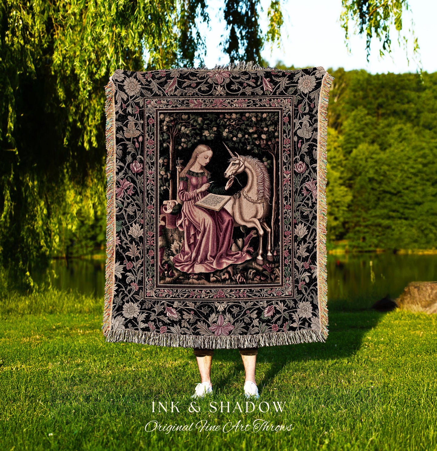 May include: A woven tapestry throw blanket with a black background and a pink and cream floral pattern. The center of the blanket features a framed image of a woman in a pink dress sitting with a unicorn. The text 'INK & SHADOW Original Fine Art Throws' is printed below the blanket.