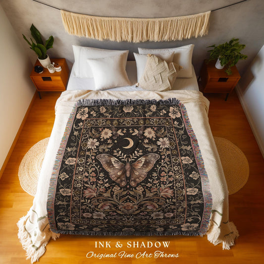 May include: A woven throw blanket features a dark background with a central design of a butterfly surrounded by flowers and a crescent moon. The design is framed by a decorative border. The throw is displayed folded on a bed.  The text 'INK & SHADOW Original Fine Art Throws' is visible at the bottom.