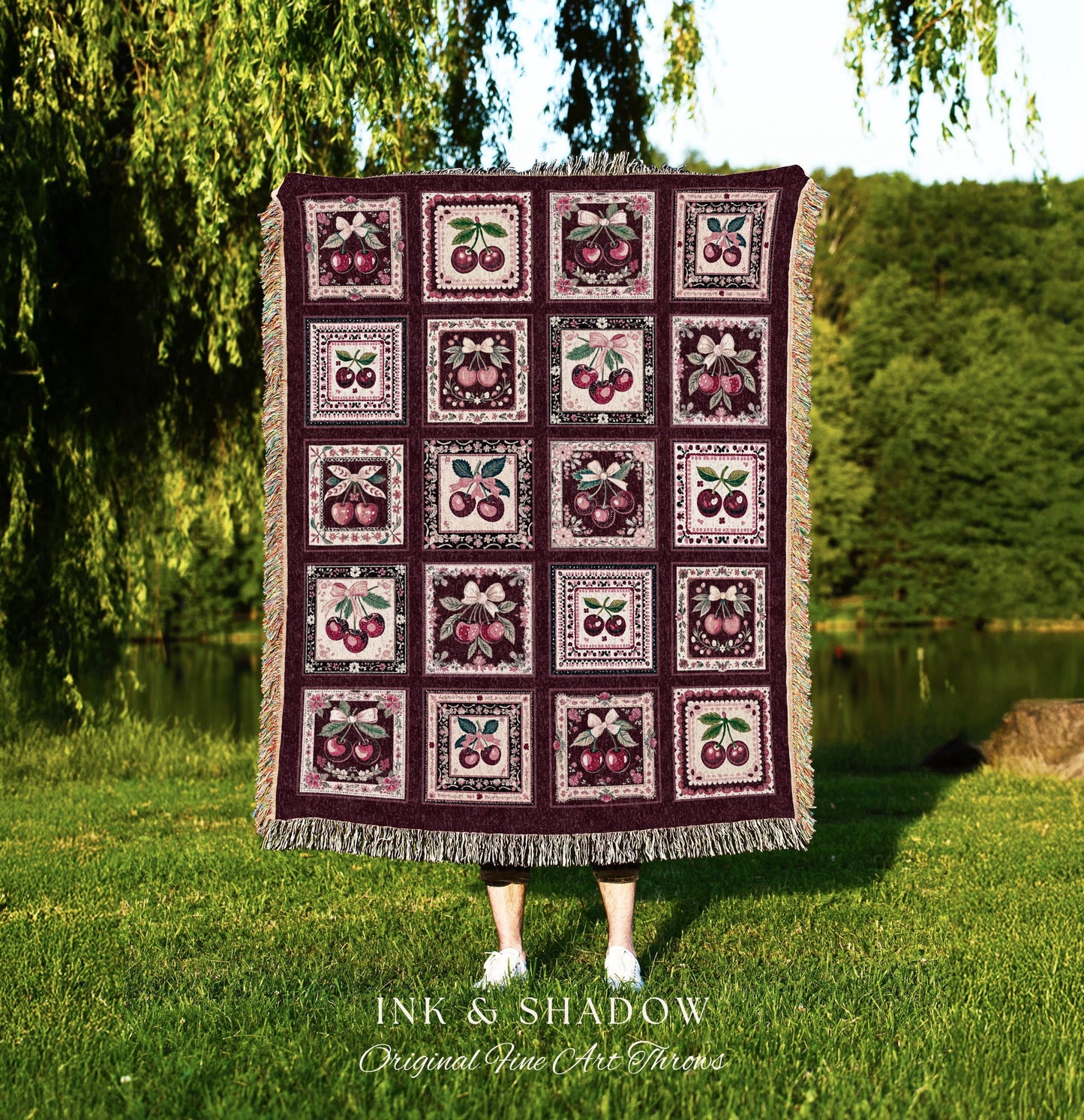 May include: A woven throw blanket with a burgundy background and a repeating pattern of framed cherries. The blanket is made of a soft, textured fabric and has a fringed edge. The text 'INK & SHADOW Original Fine Art Throws' is printed at the bottom of the blanket.