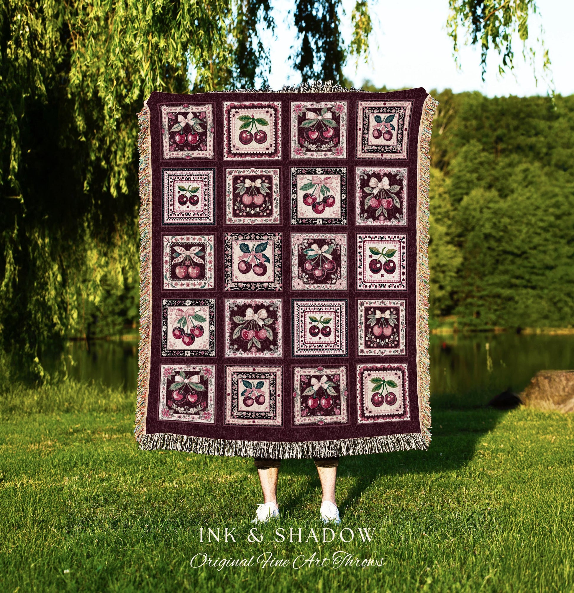 May include: A woven throw blanket with a burgundy background and a repeating pattern of framed cherries. The blanket is made of a soft, textured fabric and has a fringed edge. The text 'INK & SHADOW Original Fine Art Throws' is printed at the bottom of the blanket.