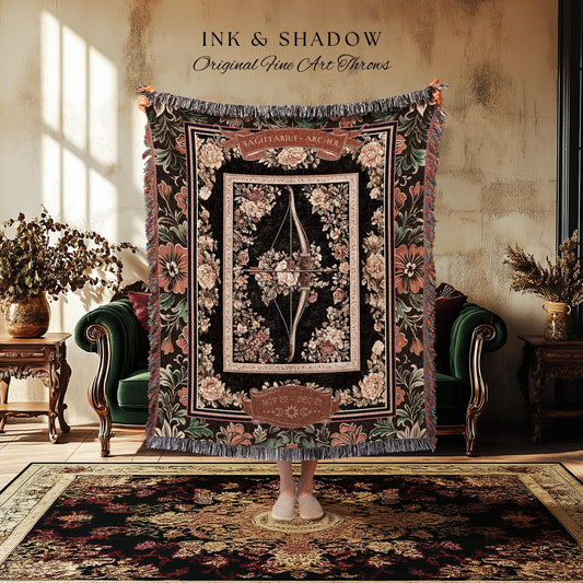 May include: A black and brown tapestry throw blanket with a floral design. The center of the blanket features a bow and arrow with the words 'Sagittarius - Archer' above it. The blanket has a fringe border.