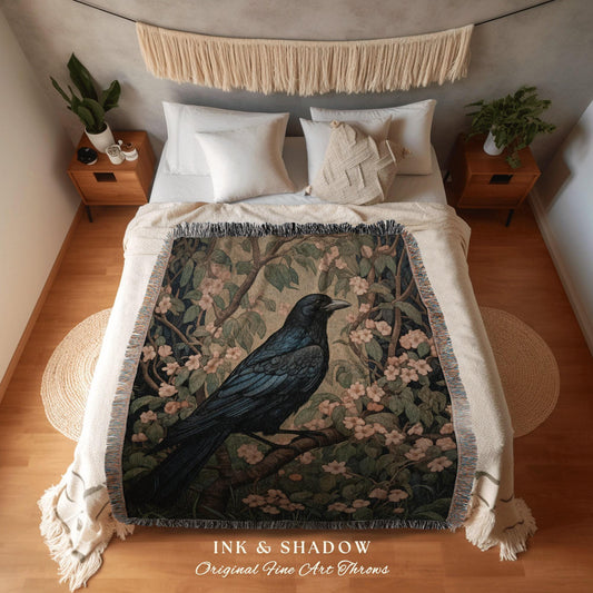 May include: A woven throw blanket with a black crow perched on a branch with pink flowers. The throw blanket has a dark green background with a light green and pink floral pattern. The throw blanket has the words 'INK & SHADOW Original Fine Art Throws' at the bottom.