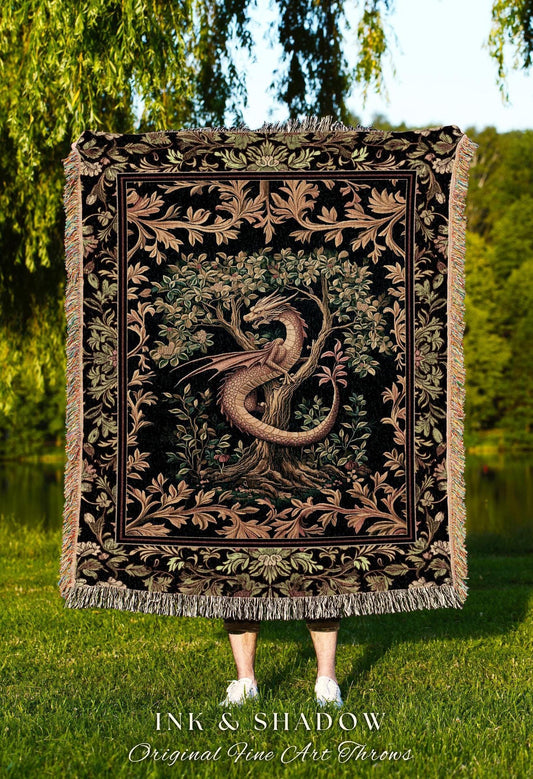 May include: A woven tapestry throw blanket features a dragon coiled around a tree in a lush garden. The design is in shades of dark brown, beige, and green on a black background.  The throw is bordered with an ornate floral pattern.  Text on the image reads: 'INK & SHADOW Original Fine Art Throws'.