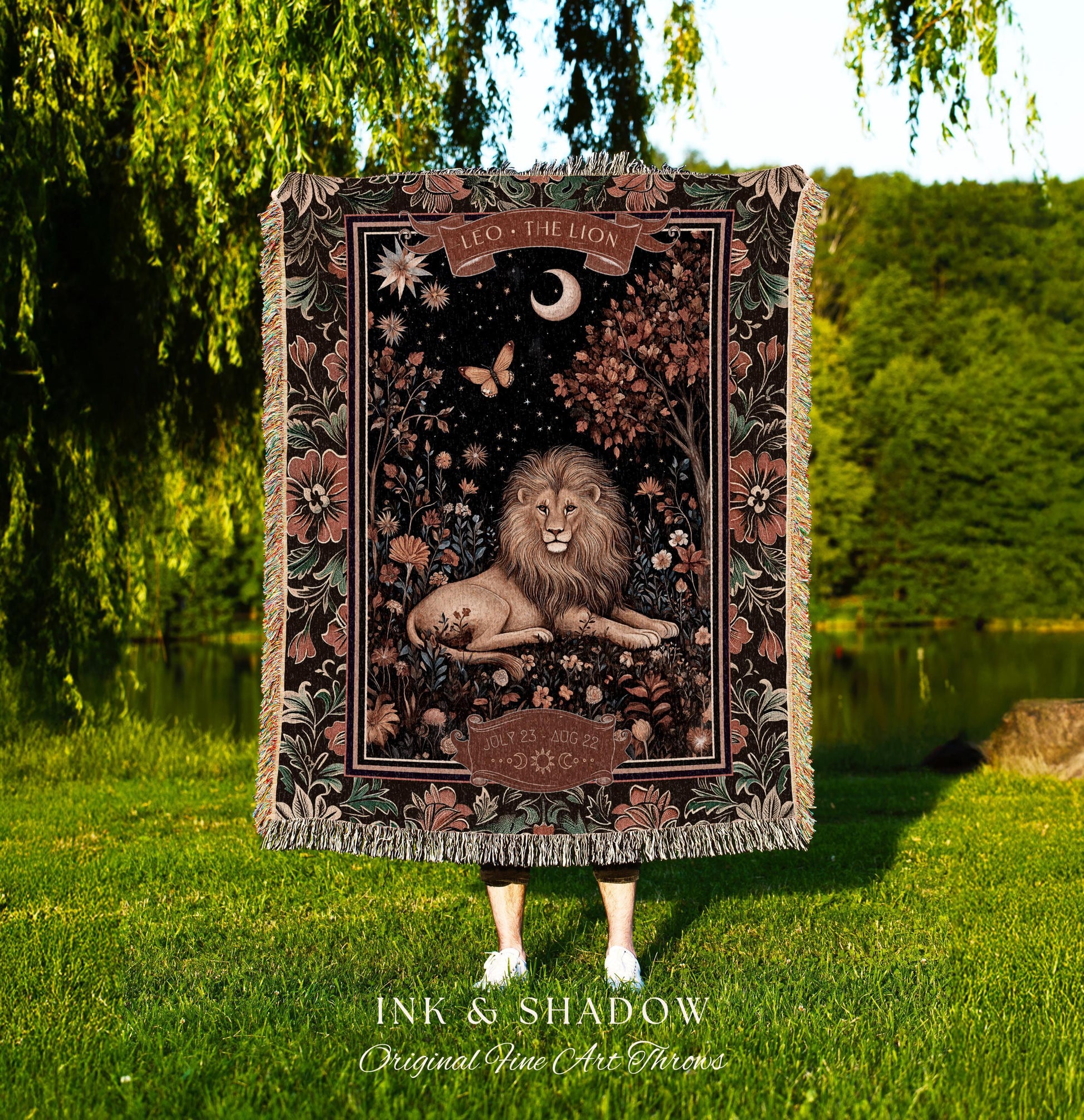 May include: A woven tapestry throw blanket with a dark background and a lion in the center. The lion is surrounded by flowers and leaves. The tapestry has a border of flowers and leaves. The text 'LEO - THE LION' is at the top of the tapestry. The text 'INK & SHADOW Original Fine Art Throws' is at the bottom of the tapestry.