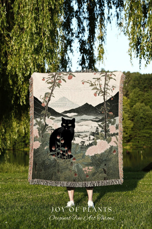 May include: A woven throw blanket featuring a black cat in a landscape scene with mountains, trees, and flowers. The blanket has a fringed edge and the text "JOY OF PLANTS Original Fine Art Throws".