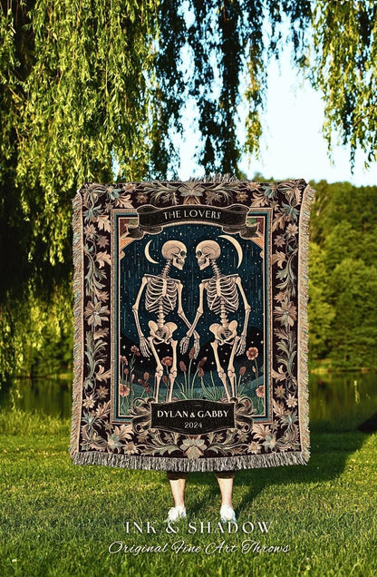 May include: A decorative throw blanket featuring a dark background with two skeletons holding hands. The words "THE LOVERS" and "DYLAN & GABBY 2024" are displayed on the blanket. The blanket has a floral border and fringe.