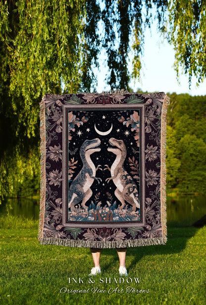 May include: A tapestry throw blanket featuring two Tyrannosaurus Rex dinosaurs facing each other under a crescent moon and stars. The blanket has a dark background with floral and leaf accents, and a fringed edge. The text "INK & SHADOW Original Fine Art Throws" is at the bottom.