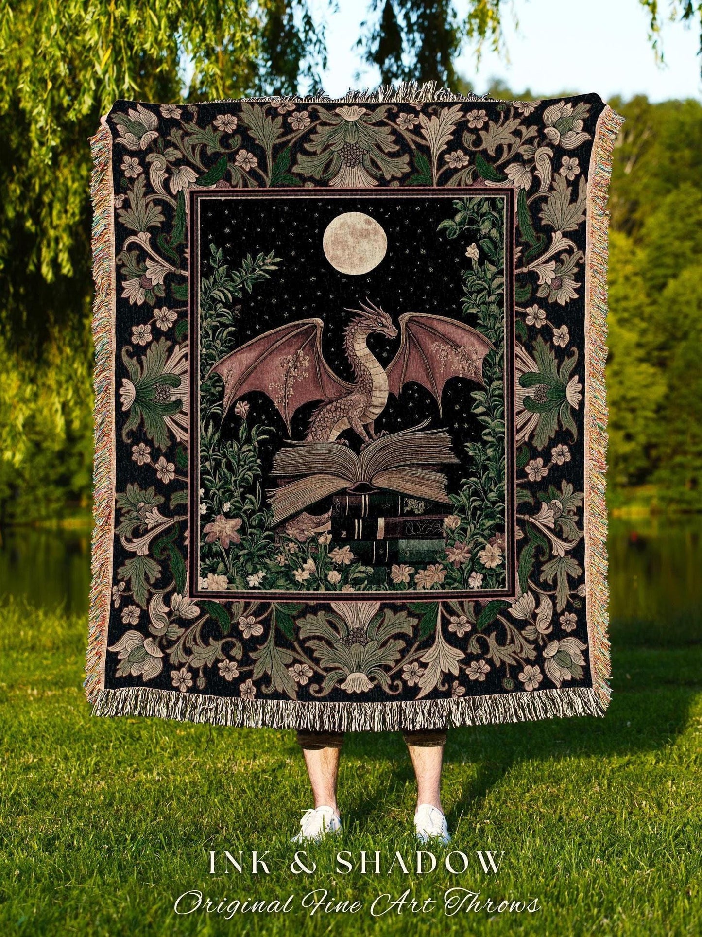 May include: A woven throw blanket featuring a dragon perched on books under a full moon and starry sky. The dragon has red-brown wings and is surrounded by floral and foliage designs. The blanket has a black background with a decorative border and fringe. The text "INK & SHADOW Original Fine Art Throws" is at the bottom.
