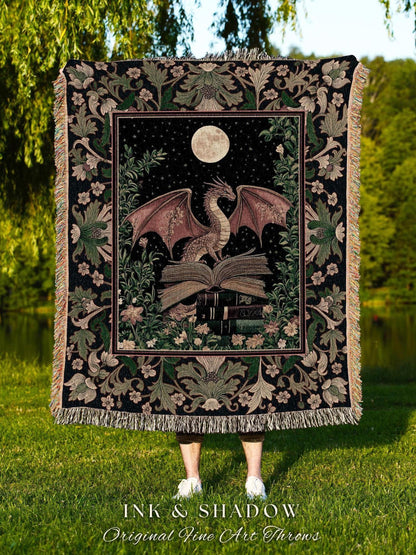 May include: A woven throw blanket featuring a dragon perched on books under a full moon and starry sky. The dragon has red-brown wings and is surrounded by floral and foliage designs. The blanket has a black background with a decorative border and fringe. The text "INK & SHADOW Original Fine Art Throws" is at the bottom.