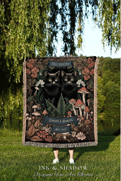 May include: A woven throw blanket featuring two black cats with yellow eyes, surrounded by mushrooms, flowers, and foliage. The blanket has the text "THE LOVERS", "LONDON & DAKOTA", and "EST. 2023". The bottom of the blanket reads "INK & SHADOW Original Fine Art Throws".