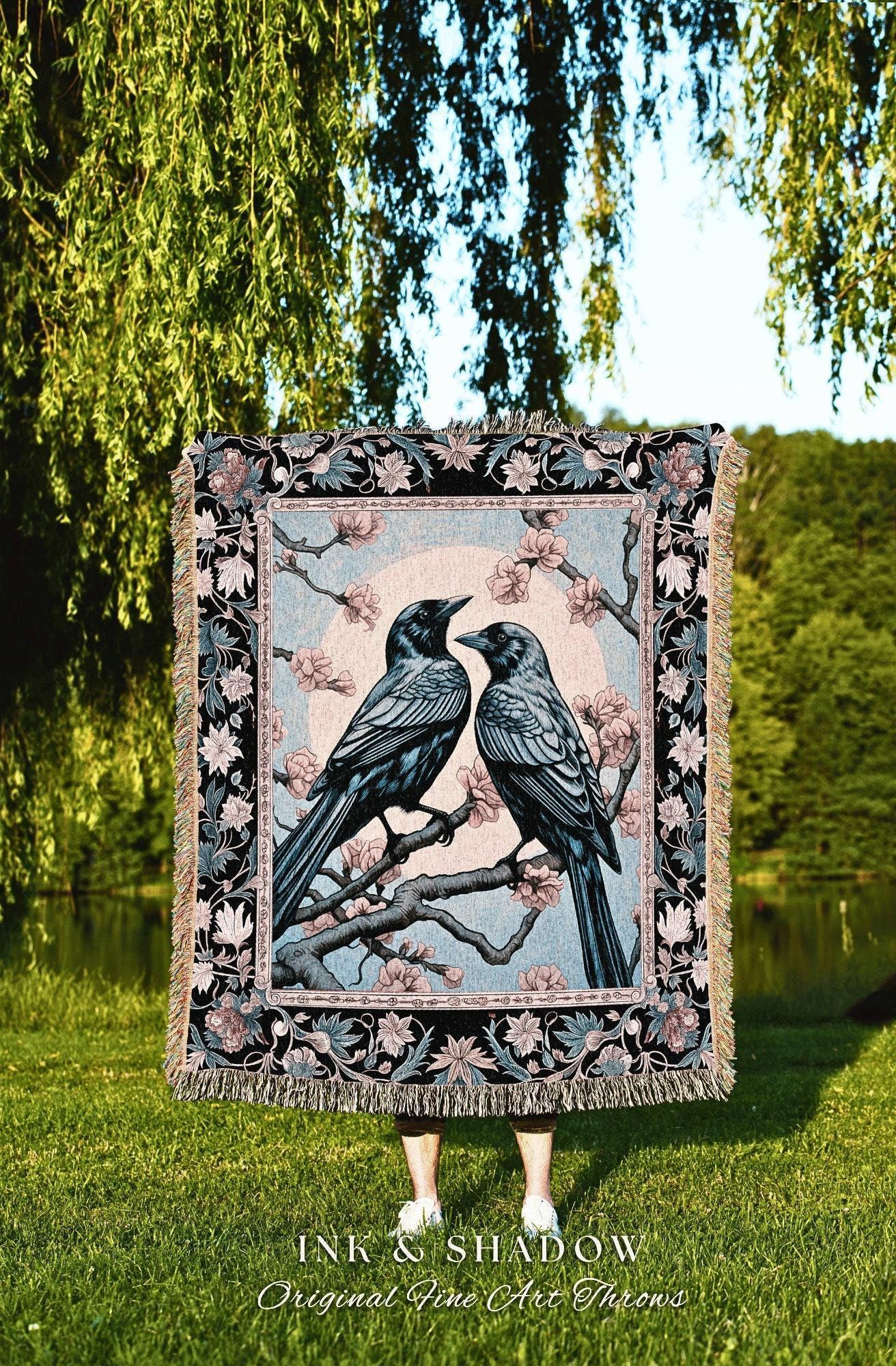 May include: A woven throw blanket featuring two crows perched on a flowering branch, set against a light blue background. The blanket has a black border with floral and leaf patterns, and a fringed edge. The text "INK & SHADOW Original Fine Art Throws" is at the bottom.