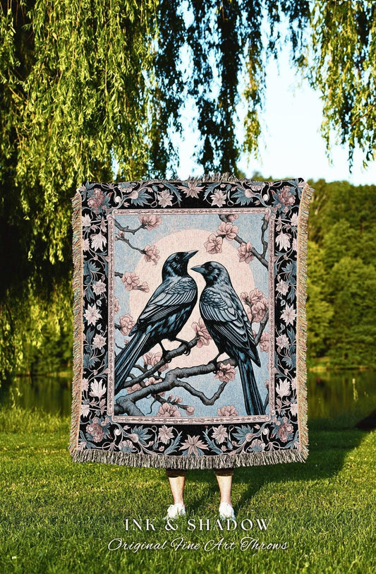 May include: A woven throw blanket featuring two crows perched on a flowering branch, set against a light blue background. The blanket has a black border with floral and leaf patterns, and a fringed edge. The text "INK & SHADOW Original Fine Art Throws" is at the bottom.