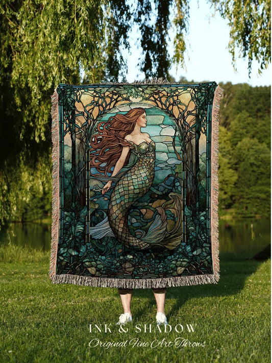 May include: A woven throw blanket featuring a mermaid illustration in shades of green, blue, and brown. The artwork is framed by stylized trees and a stained-glass effect. The blanket has a fringed edge and the text "INK & SHADOW Original Fine Art Throws".