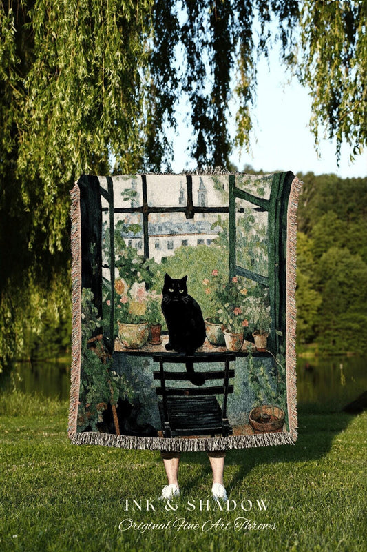 May include: A tapestry throw featuring a black cat sitting on a small table in front of a window with a garden scene. The throw has a fringed edge and the text "INK & SHADOW Original Fine Art Throws".