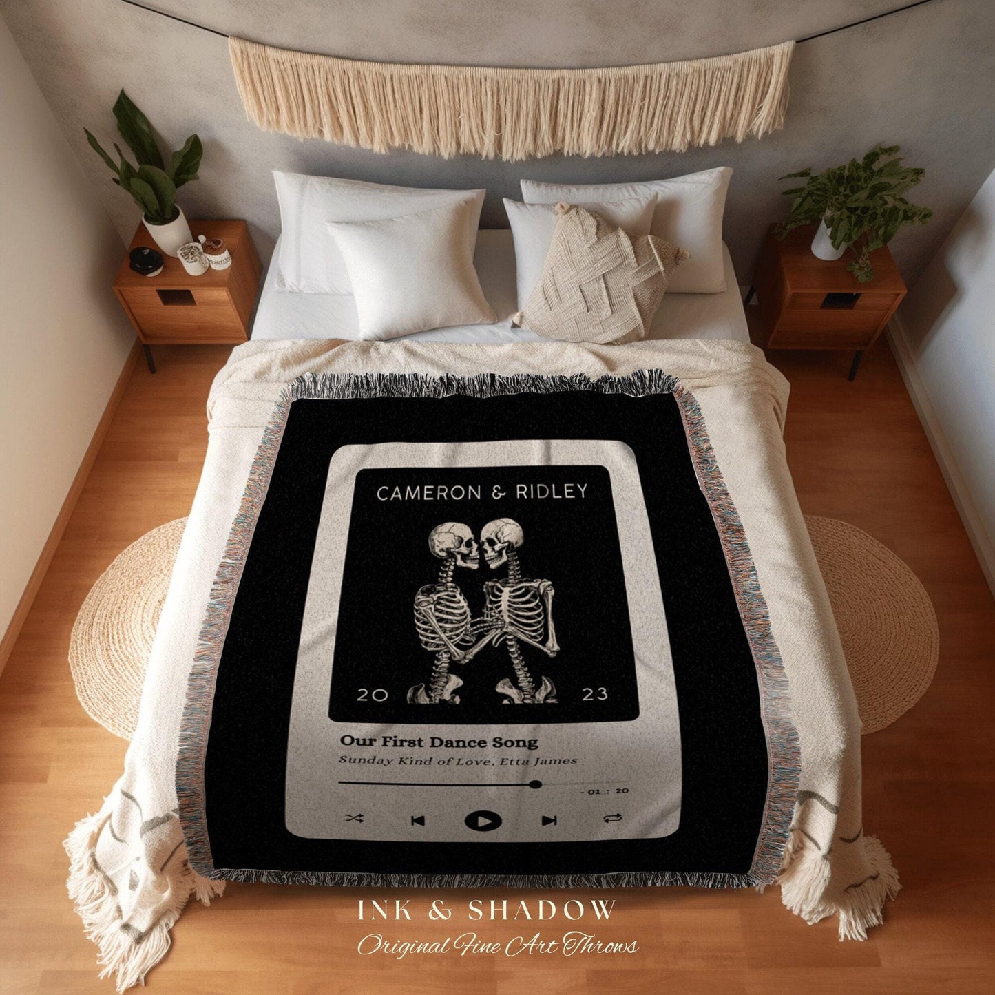 May include: A black throw blanket with a white border and fringe. The blanket features a graphic of two skeletons embracing, with text that reads "CAMERON & RIDLEY" and "Our First Dance Song". The blanket is on a bed with white pillows.