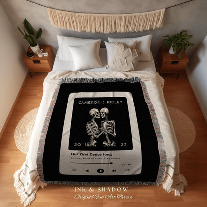 May include: A black throw blanket with a white border and fringe. The blanket features a graphic of two skeletons embracing, with text that reads "CAMERON & RIDLEY" and "Our First Dance Song". The blanket is on a bed with white pillows.