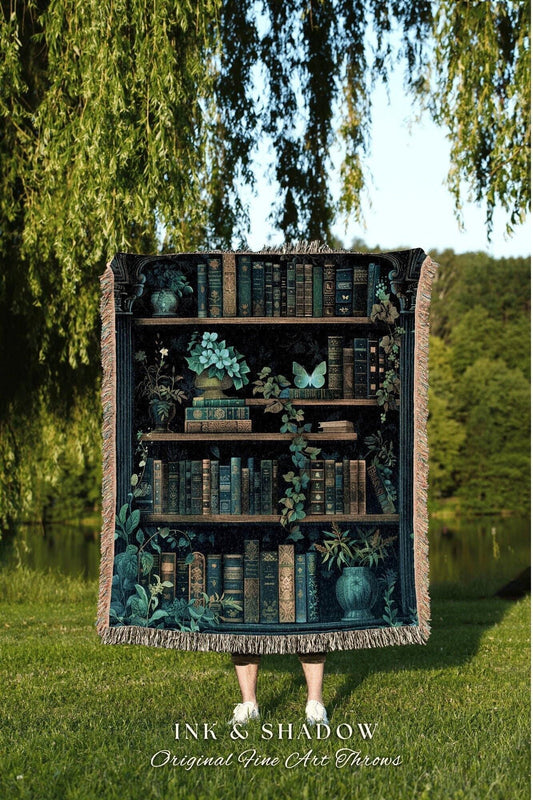 May include: A tapestry throw blanket featuring a detailed illustration of a bookshelf filled with books, plants, and a butterfly. The throw has a dark teal and brown color scheme with a fringed edge. The text "INK & SHADOW Original Fine Art Throws" is at the bottom.