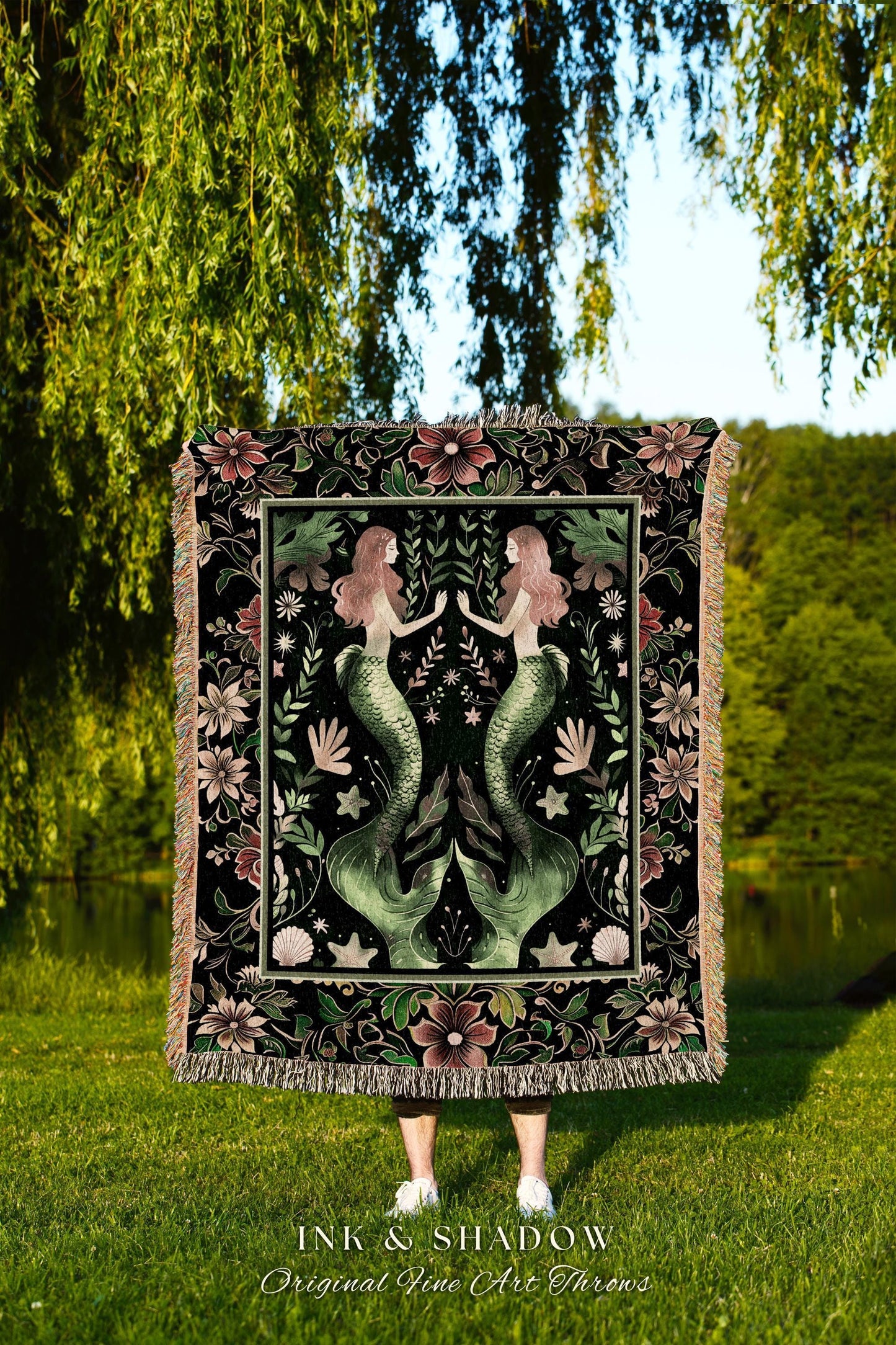May include: A woven tapestry throw blanket with a dark background featuring two mermaids with green tails and pink flowers. The text "INK & SHADOW Original Fine Art Throws" is at the bottom of the blanket.