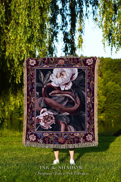 May include: A decorative throw blanket featuring a floral and snake design. The blanket has a dark burgundy border with a cream-colored fringe. The artwork depicts a snake coiled around white and pink flowers. The text "INK & SHADOW Original Fine Art Throws" is at the bottom.