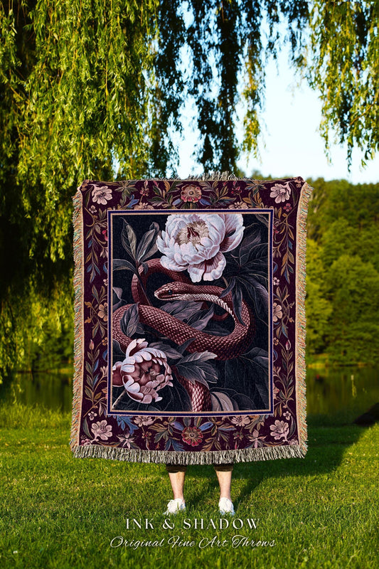 May include: A decorative throw blanket featuring a floral and snake design. The blanket has a dark burgundy border with a cream-colored fringe. The artwork depicts a snake coiled around white and pink flowers. The text "INK & SHADOW Original Fine Art Throws" is at the bottom.