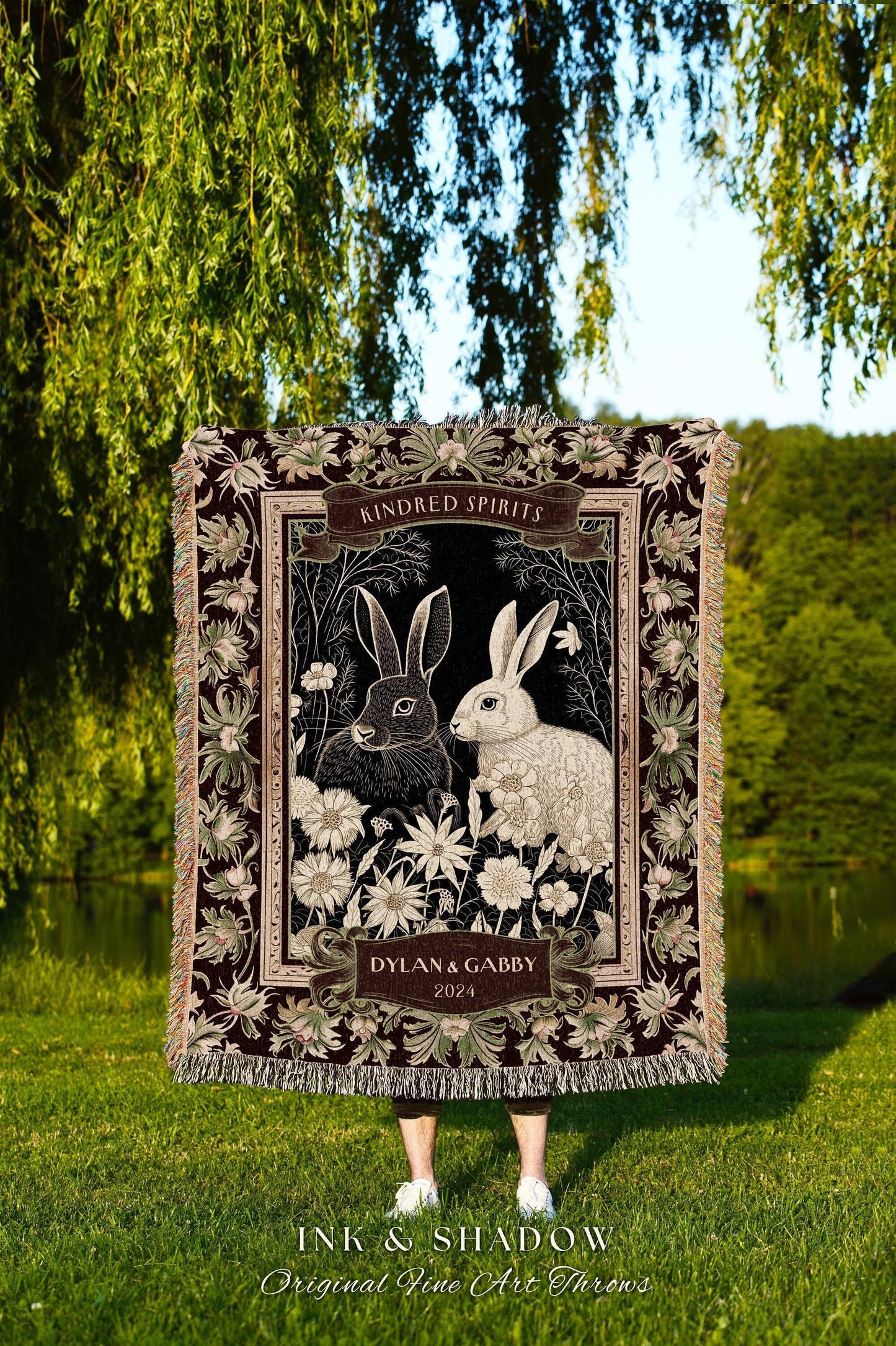 May include: A decorative throw blanket featuring a black and white illustration of two rabbits surrounded by flowers. The blanket has a dark burgundy border with floral accents and the text "Kindred Spirits" and "Dylan & Gabby 2024". The blanket is fringed.
