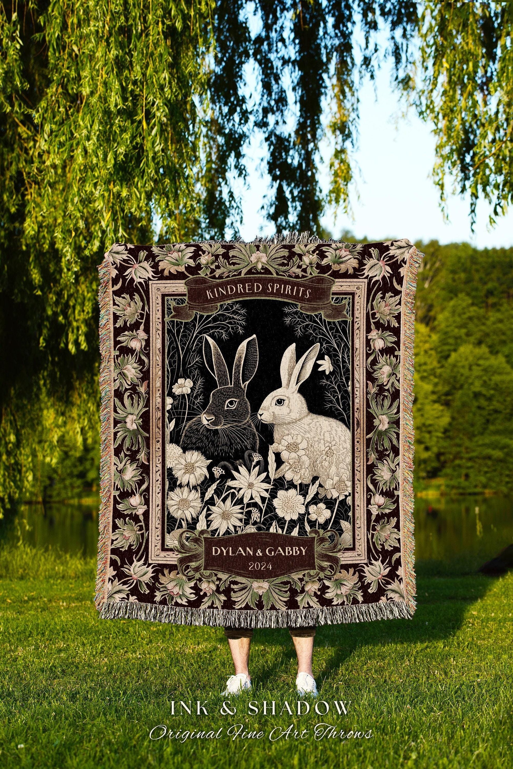 May include: A decorative throw blanket featuring a black and white illustration of two rabbits surrounded by flowers. The blanket has a dark burgundy border with floral accents and the text "Kindred Spirits" and "Dylan & Gabby 2024". The blanket is fringed.