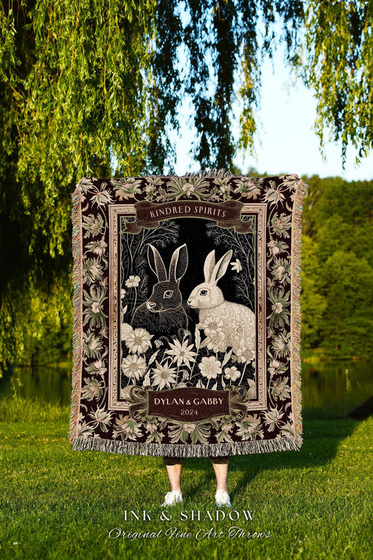 May include: A decorative throw blanket featuring a black and white illustration of two rabbits surrounded by flowers. The blanket has a dark burgundy border with floral accents and the text "Kindred Spirits" and "Dylan & Gabby 2024". The blanket is fringed.