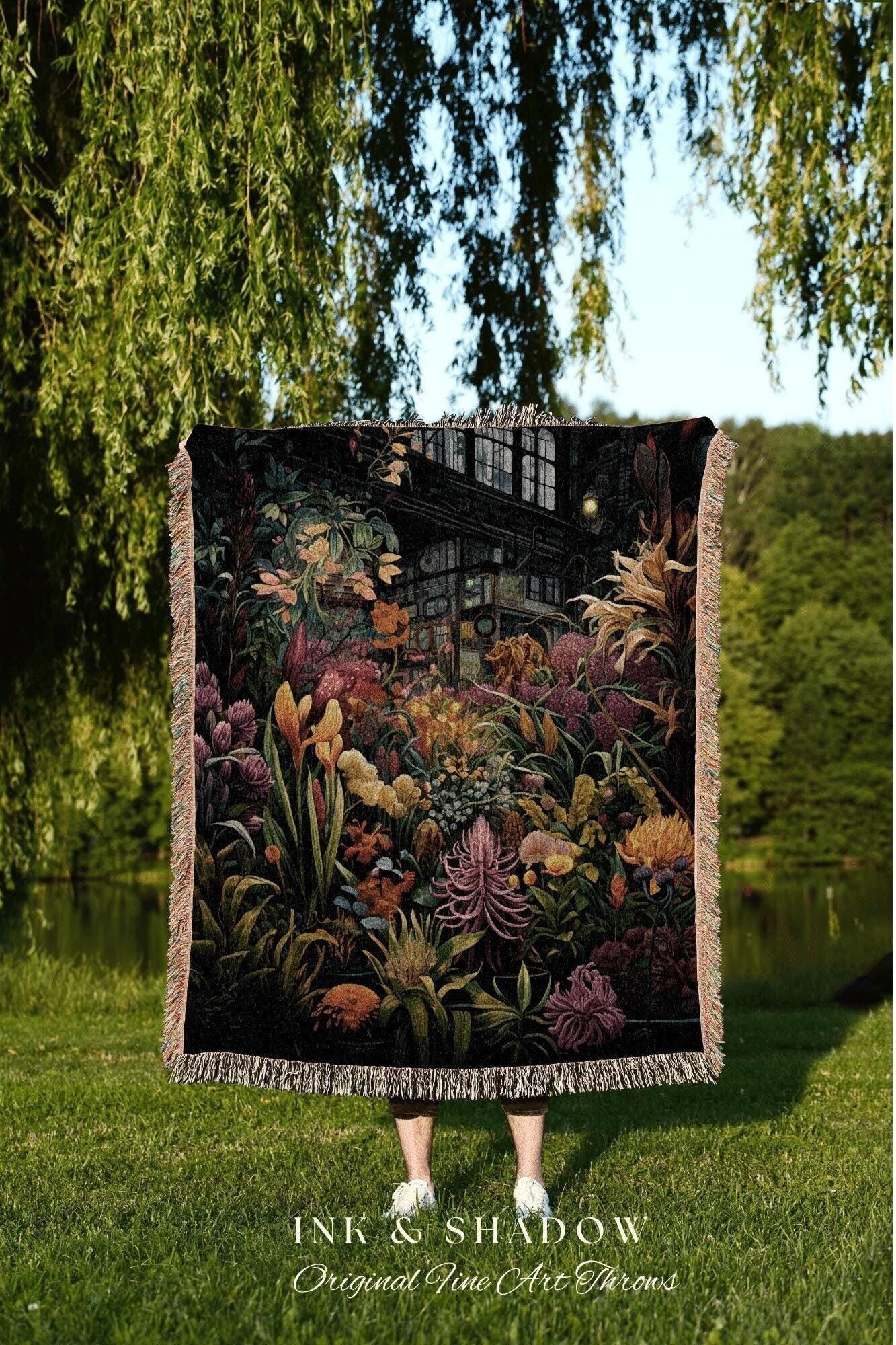 May include: A woven throw blanket featuring a detailed botanical illustration with a greenhouse in the background. The artwork showcases a variety of colorful flowers and plants in shades of pink, yellow, and green. The blanket has a fringed edge and the text "INK & SHADOW Original Fine Art Throws".