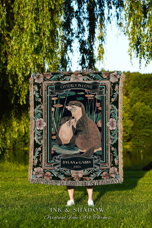May include: A decorative throw blanket featuring an illustration of two otters in a pond, with the text "OTTERLY IN LOVE" and "DYLAN & GABBY 2024". The blanket has a dark green background with floral and leaf accents and a fringed edge.