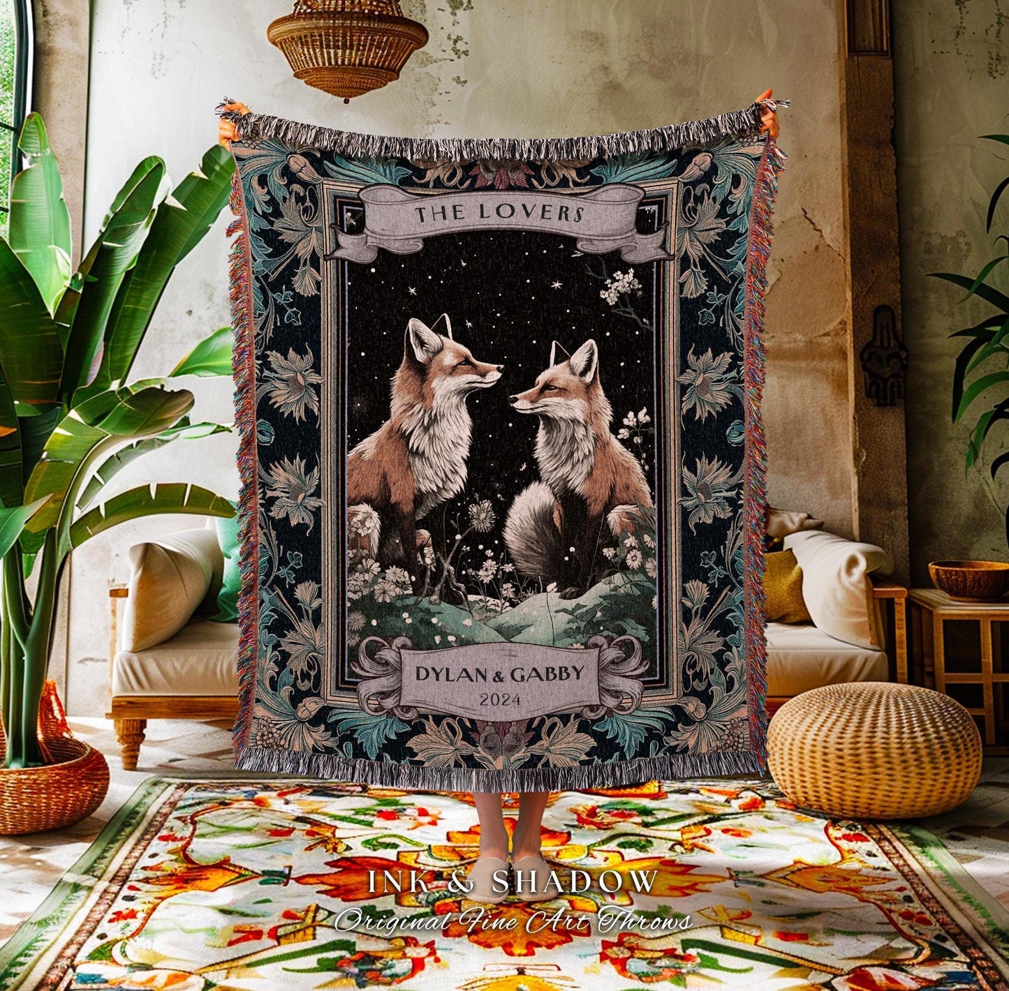 May include: A woven throw blanket featuring two foxes facing each other under a starry night sky. The blanket has a dark background with floral and leaf patterns, and the text "THE LOVERS" and "DYLAN & GABBY 2024".