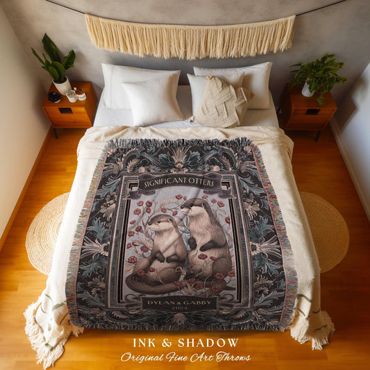 May include: A woven throw blanket featuring a detailed illustration of two otters with the text "Significant Otters" and "Dylan & Gabby 2023". The blanket has a dark floral border and fringe.