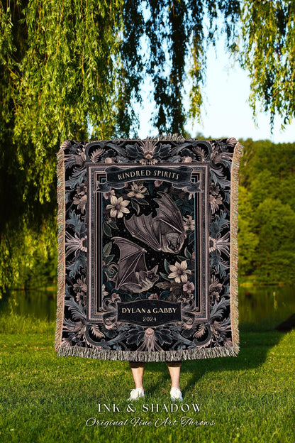 May include: A black throw blanket with a floral and bat design. The blanket features the text "Kindred Spirits" and "Dylan & Gabby 2024". The bottom of the blanket has the text "INK & SHADOW Original Fine Art Throws".