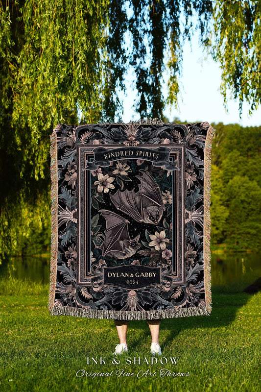 May include: A black throw blanket with a floral and bat design. The blanket features the text "Kindred Spirits" and "Dylan & Gabby 2024". The bottom of the blanket has the text "INK & SHADOW Original Fine Art Throws".