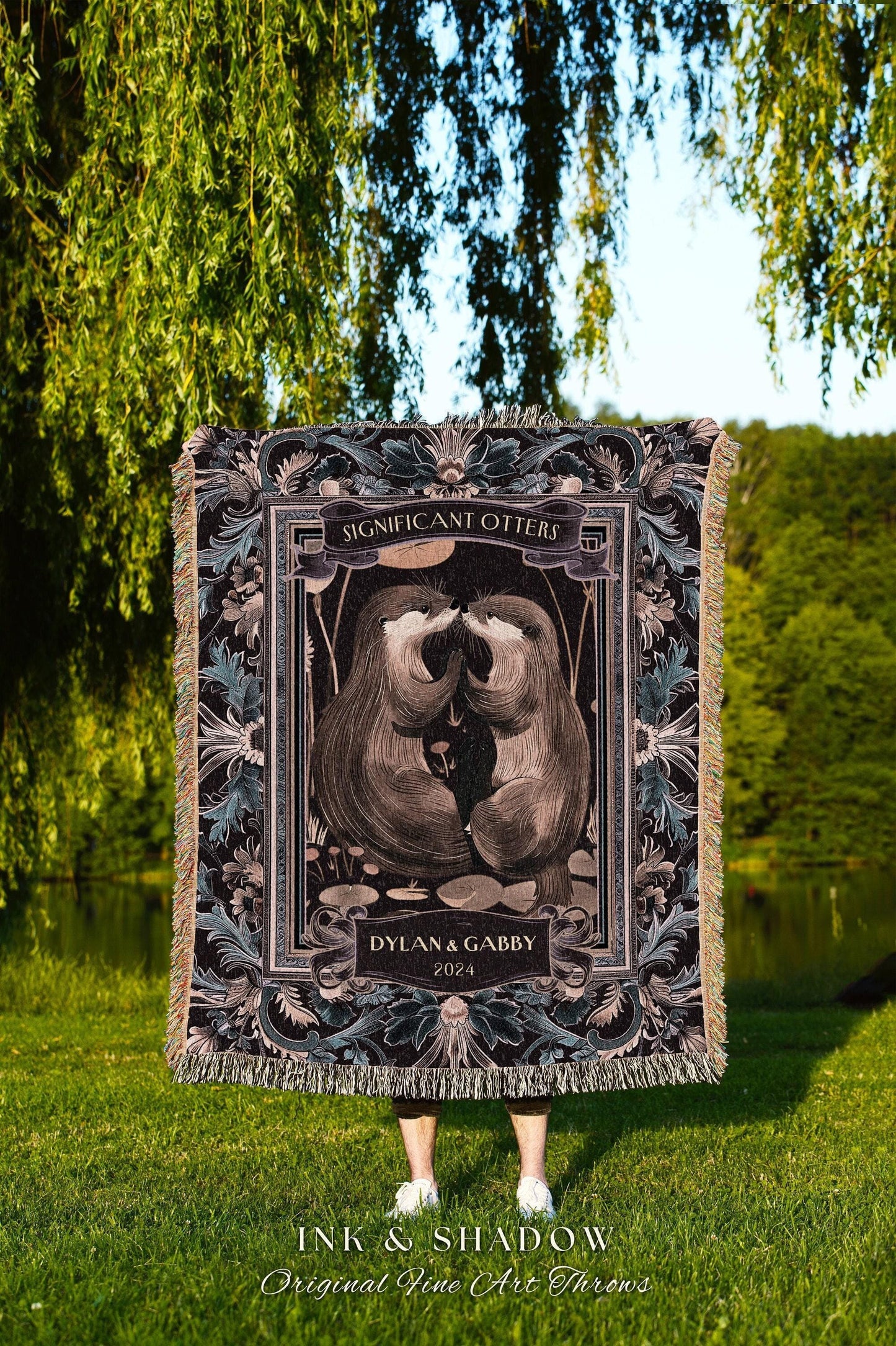 May include: A woven throw blanket featuring a central illustration of two otters touching noses, framed by floral and geometric patterns. The text "Significant Otters" and "Dylan & Gabby 2024" are displayed. The blanket has a fringed edge.