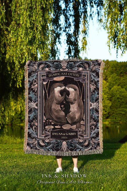 May include: A woven throw blanket featuring a central illustration of two otters touching noses, framed by floral and geometric patterns. The text "Significant Otters" and "Dylan & Gabby 2024" are displayed. The blanket has a fringed edge.