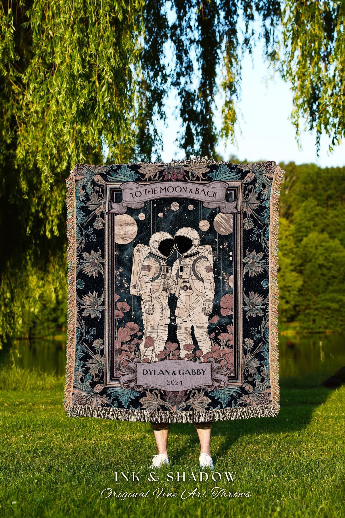 May include: A woven throw blanket featuring a design of two astronauts in space suits, with the text "TO THE MOON & BACK" at the top. The blanket includes the names "DYLAN & GABBY" and the year "2024". The bottom of the blanket has the text "INK & SHADOW Original Fine Art Throws".