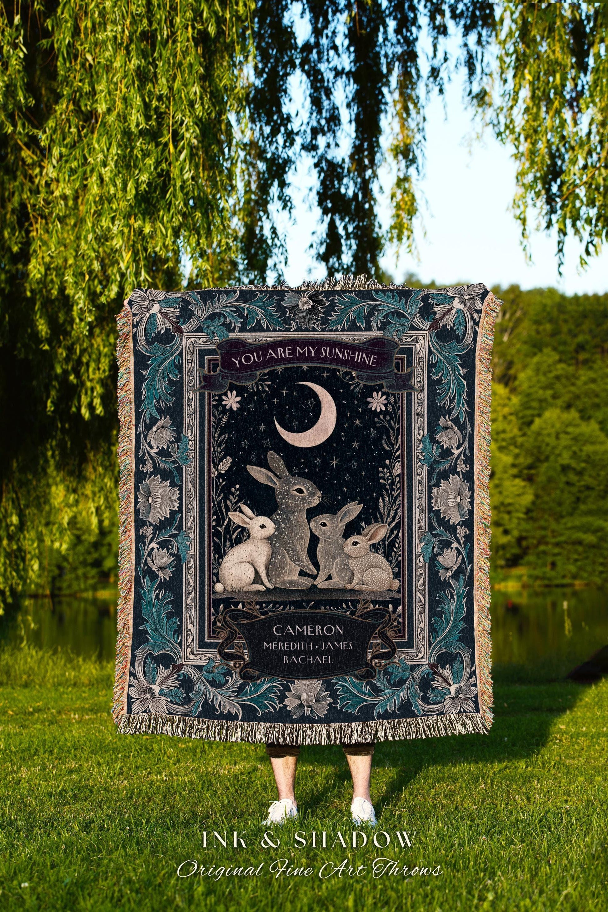 May include: A dark teal throw blanket with a detailed illustration of rabbits under a crescent moon. The blanket has a floral border and the text "YOU ARE MY SUNSHINE" and "INK & SHADOW Original Fine Art Throws".