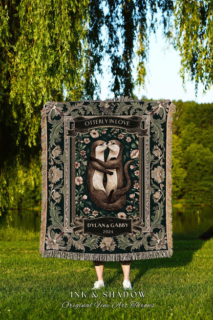 May include: A dark green throw blanket featuring a romantic illustration of two otters embracing, with the text "OTTERLY IN LOVE" and "DYLAN & GABBY 2024". The blanket has a decorative floral border and fringe.