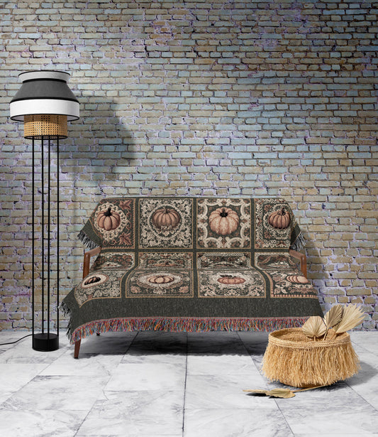 May include: A decorative throw blanket with a pumpkin pattern draped over a wooden sofa. The blanket features a repeating design of pumpkins in squares, with a dark green border and fringe. A tall black floor lamp and a woven basket are also in the image.
